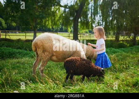 Ein bezauberndes kleines Mädchen, das auf dem Bauernhof mit Ziegen und Schafen spielt. Kind, das sich mit Tieren vertraut macht. Landwirtschaft und Gartenarbeit für Kleinkinder. Im Freien Stockfoto