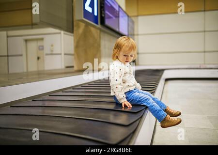 Ein bezauberndes kleines Mädchen am Flughafen. Das Kleinkind sitzt auf dem Gepäckkarussell und wartet auf sein Gepäck. Reisen mit kleinen Kindern Stockfoto