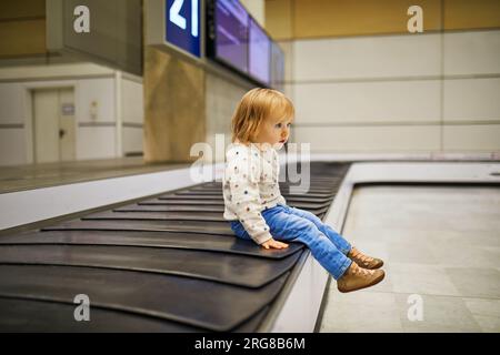 Ein bezauberndes kleines Mädchen am Flughafen. Das Kleinkind sitzt auf dem Gepäckkarussell und wartet auf sein Gepäck. Reisen mit kleinen Kindern Stockfoto