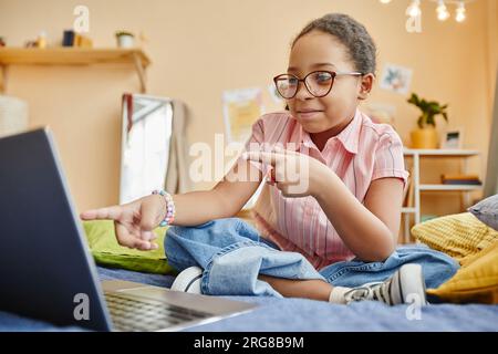 Süßes Schulmädchen, das auf dem Bett sitzt und während der Online-Kommunikation in einem Video-Chat oder während eines Fernstudiums auf den Laptop-Bildschirm zeigt Stockfoto