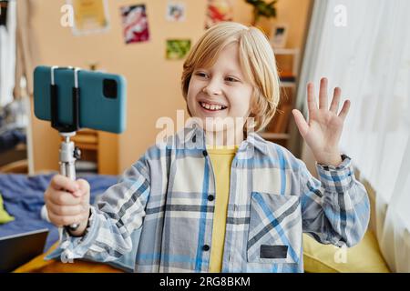 Blonder, lächelnder Schuljunge und Blogger winken dem Online-Publikum die Hand, während sie Livestream machen und auf den Smartphone-Bildschirm schauen Stockfoto