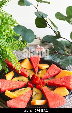 Scheiben von Wassermelonen, Cantaloupe, Kiwi und Erdbeeren werden auf einem schwarzen Teller angeordnet, um einen lebendigen Obstsalat zu kreieren, der perfekt für einen erfrischenden Sommer ist Stockfoto