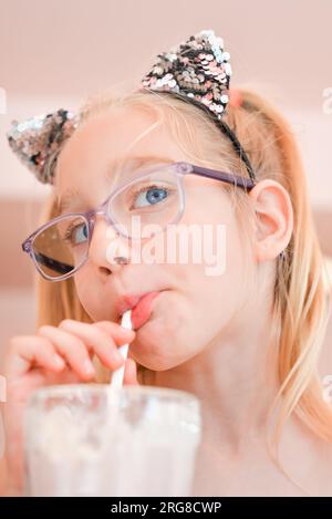 Ein hübsches Mädchen, sieben Jahre alt, trinkt Erdbeermilchshake durch einen Strohhalm in einem Café. Portrait des glücklichen Mädchens, das einen Cocktail trinkt Stockfoto