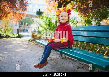 Ein bezauberndes kleines Mädchen im roten Poncho, das an einem sonnigen Herbsttag im Herbstpark spaziert. Fröhliches Kind, das sich draußen in einer Straße von Paris, Frankreich, amüsiert. Außenbereich A Stockfoto