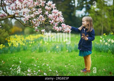 Ein bezauberndes Vorschulmädchen genießt einen schönen Frühlingstag im Park während der Magnolienblüte. Aktivitäten im Freien im Frühling für Kinder. Kleine Kinder-Entdeckungsreise Stockfoto