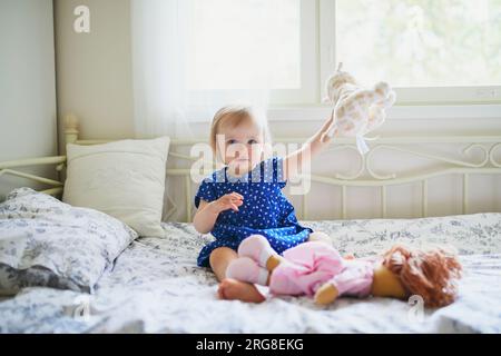 Ein niedliches kleines Mädchen in blauem Kleid, das auf dem Bett sitzt und mit Puppe, Teddybär und Hund spielt. Das kleine Kind hat Spaß im Schlafzimmer. Ein Kind, das Plüschspielzeug spielt Stockfoto