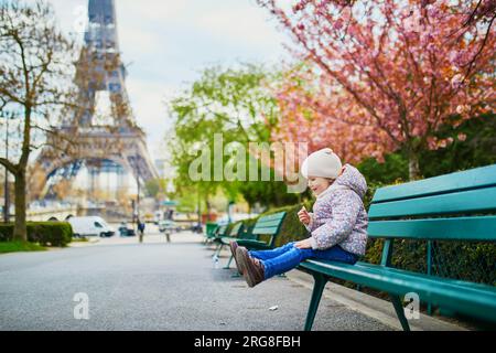 Ein bezauberndes dreijähriges Mädchen, das auf der Bank neben dem Eiffelturm in Paris, Frankreich, sitzt. Kirschblüten blühen an einem Frühlingstag in Paris in voller Blüte Stockfoto