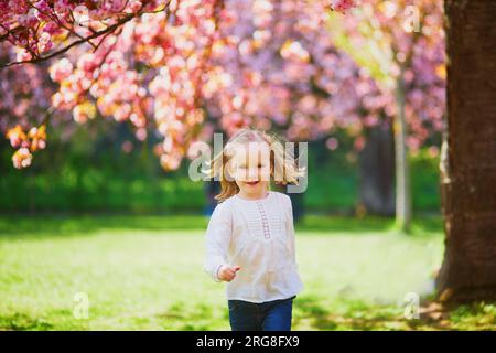 Ein bezauberndes dreijähriges Mädchen, das den sonnigen Frühlingstag im Park of Sceaux bei Paris, Frankreich, genießt. Ein wunderschönes Kind, das Spaß unter einem rosa Kirschblütenbaum hat Stockfoto