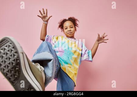 Fröhliches, verspieltes Mädchen in Freizeitkleidung, das vor der Kamera auf pinkfarbenem Hintergrund posiert und während der Fotoaufnahme an der Wand steht Stockfoto