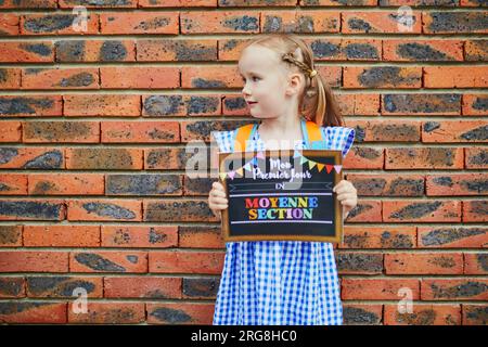 Fröhliches 4-jähriges Mädchen mit einem Plakat mit dem Text "Mein erster Tag im Kindergarten" in französischer Sprache. Bildungssystem in Frankreich und Schulanfang c Stockfoto