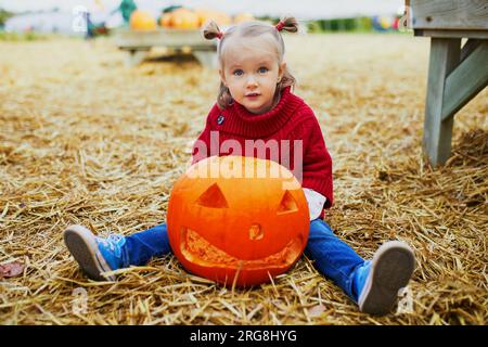 Ein niedliches Kleinkind in rotem Poncho, das auf dem Boden sitzt und auf dem Bauernhof mit dem Kürbis Jack-o'lantern spielt. Fröhliches Kind feiert Halloween Stockfoto