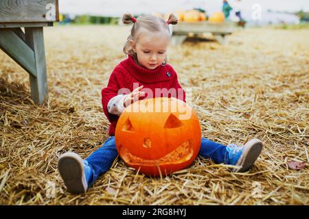 Ein niedliches Kleinkind in rotem Poncho, das auf dem Boden sitzt und auf dem Bauernhof mit dem Kürbis Jack-o'lantern spielt. Fröhliches Kind feiert Halloween Stockfoto