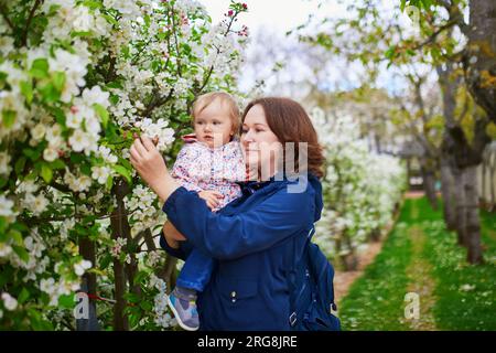 Junge Frau hält ihr 1-jähriges Mädchen an einem Frühlingstag im Park. Mutter und Tochter genießen die Apfelblütensaison. Familienaktivitäten mit k Stockfoto