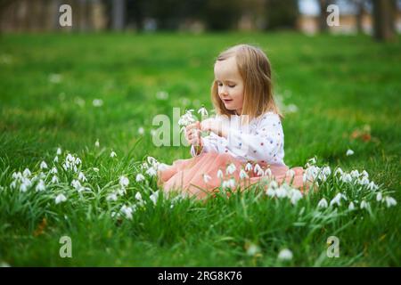 Süßes Vorschulmädchen in pinkfarbenem Tutu-Rock, das im Gras sitzt und an einem Frühlingstag im Park oder Wald viele Schneetropfen blüht. Kleiner Junge, der Natur erforscht Stockfoto