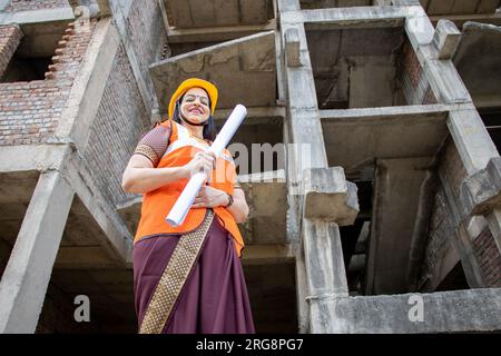 Porträt einer selbstbewussten jungen, wunderschönen indischen Bauingenieurin oder Architektin, die Helm und Weste trägt und einen Papierentwurf auf der Baustelle si hält Stockfoto