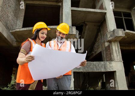 Zwei indische Bauingenieure oder Architekten mit Helm und Weste, die einen Papierkram auf der Baustelle halten, um über die richtige Lage zu sprechen Stockfoto