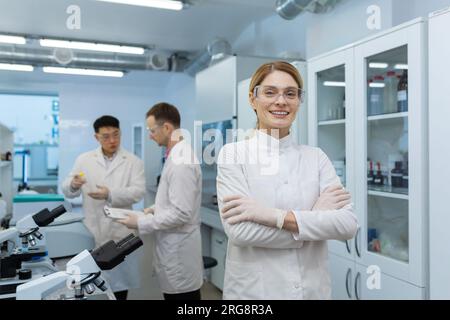 Porträt einer jungen Wissenschaftlerin, Chemikerin, Ärztin, die im Labor der Klinik steht, ihre Arme auf der Brust kreuzt und in die Kamera lächelt. Im Hintergrund, männliche Kollegen. Stockfoto