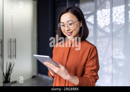 Lächelnde junge asiatische Programmiererin, die im Büro steht und an einem Tablet arbeitet. Stockfoto