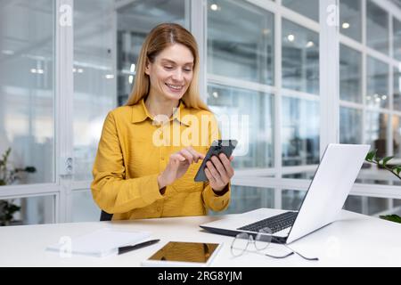 Erfahrene Geschäftsfrau im Büro am Arbeitsplatz, Mitarbeiter am Schreibtisch, die lächelnd das Telefon in der Hand halten, Chef, der die Anwendung auf dem Smartphone verwendet, Internetseiten durchsucht. Stockfoto