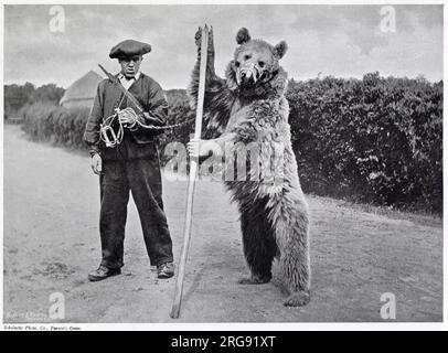 Junge Wildbären wurden sowohl von Franzosen auf der französischen Seite der Berge als auch von Spaniern gefangen, um Tricks zur Unterhaltung zu machen. Das Foto zeigt einen Bergsteiger mit roter Kappe und einem Bären mit einem kleinen Horn und Sträuchern, von dem der Bär tanzen und seinen Stock wie eine Waffe Schultern muss. Stockfoto