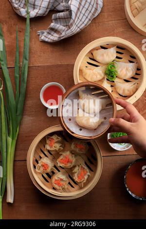 Draufsicht verschiedene Dimsum auf Bamboo Dampfgarer. Weibliche Hand legt Bambusdampfer auf den Tisch Stockfoto
