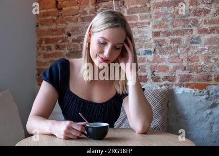 Eine junge blonde Frau sitzt an einem Tisch in einem altmodischen Café in der Stadt. Sie hält einen Kaffee in der einen Hand, die andere Hand in ihrem Haar und legt ihren Kopf in einen bedächtigen, leicht beunruhigten Ausdruck. Nachdenkliche blonde Frau im Urban Coffee Shop. Hochwertige Fotos Stockfoto