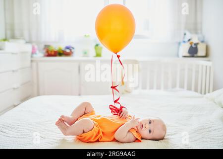 Ein bezauberndes Mädchen im orangefarbenen Strampelanzug liegt im Kinderzimmer mit farbenfrohem Ballon auf dem Bett Stockfoto