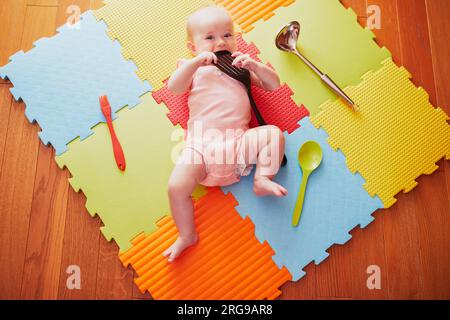 Ein kleines Mädchen mit Küchenutensilien. Das kleine Kind liegt auf der Spielmatte und spielt mit Küchenutensilien. Echte Dinge als Kinderspielzeug. Säuglingskind in der sonnigen Krankenschwester Stockfoto