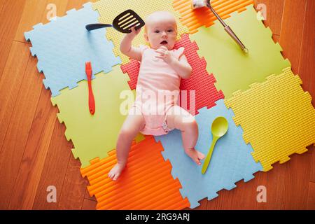 Ein kleines Mädchen mit Küchenutensilien. Das kleine Kind liegt auf der Spielmatte und spielt mit Küchenutensilien. Echte Dinge als Kinderspielzeug. Säuglingskind in der sonnigen Krankenschwester Stockfoto