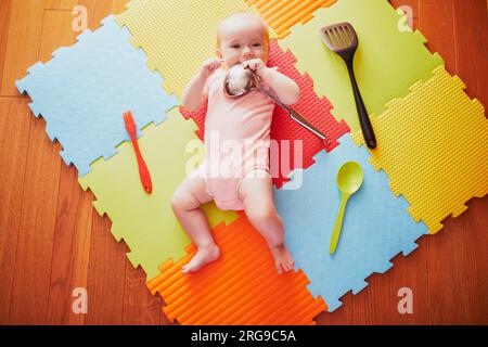 Ein kleines Mädchen mit Küchenutensilien. Das kleine Kind liegt auf der Spielmatte und spielt mit Küchenutensilien. Echte Dinge als Kinderspielzeug. Säuglingskind in der sonnigen Krankenschwester Stockfoto