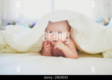 Süßes kleines Mädchen, das auf dem Bett spielt. Glückliches Kind, das Spaß im Kinderzimmer hat. Baby versteckt sich unter der Decke Stockfoto