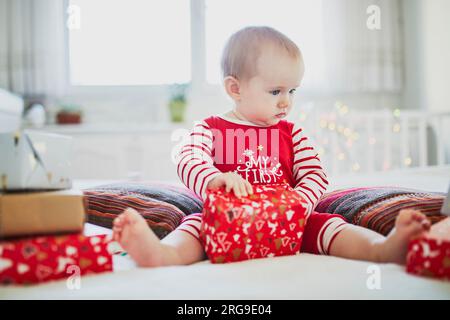 Ein glückliches kleines Mädchen, das einen Schlafanzug trägt und Weihnachtsgeschenke an ihrem ersten Weihnachtsfest eröffnete. Weihnachten mit Kindern zu Hause feiern Stockfoto