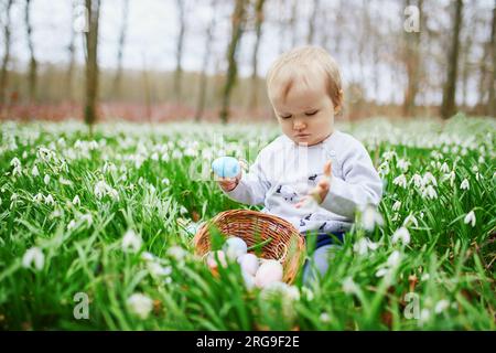 Süßes, kleines, einjähriges Mädchen, das Ostern auf Eierjagd spielt. Das Kleinkind sitzt auf dem Rasen mit vielen Schneetropfen und sammelt farbenfrohe Eier im Baske Stockfoto