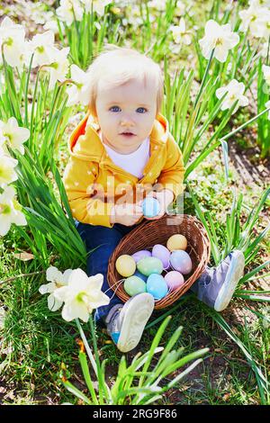 Ein einjähriges Mädchen spielt Ostern auf Eierjagd. Das Kleinkind sitzt auf dem Rasen mit vielen Narzissen und sammelt bunte Eier im Korb. Kleiner Kinderfreund Stockfoto