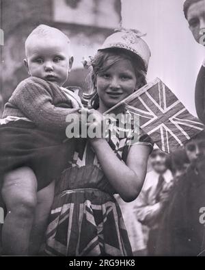 VE-Tag. Ein kleines Mädchen, patriotisch gekleidet in einem Union-Jack-Kleid, hilft einem dicken Baby, eine Flagge zu schwenken. Stockfoto