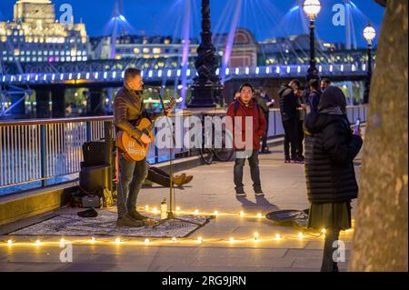 LONDON - 22. April 2023: Abendunterhaltung: Busker verzaubert Touristen in Southbank, London, unter warmem Licht vor einem dunkelblauen Abendhimmel. Stockfoto