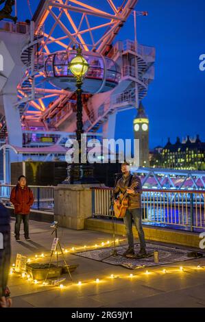 LONDON - 22. April 2023: Bezaubernde Nacht: Busker unterhält Touristen am London Eye auf der Southbank, warme Lichter vor tiefblauem Himmel, mit Big Ben Stockfoto