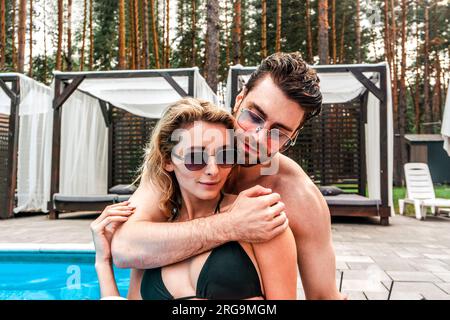 Ein verliebter Mann umarmt seine Freundin in einer Sonnenbrille, während er hinter ihr am Pool sitzt. Romantik- und Sommerurlaubskonzept Stockfoto