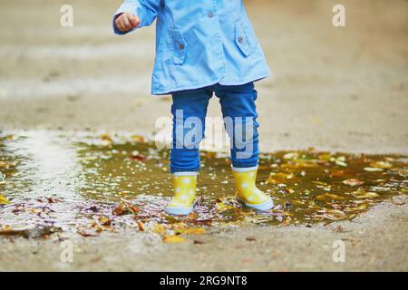 Ein Kind trägt gelbe Regenstiefel und springt an einem Herbsttag in die Pfütze. Ein kleines Mädchen hat Spaß mit Wasser und Schlamm im Park an einem regnerischen Tag. Herbst im Freien Stockfoto