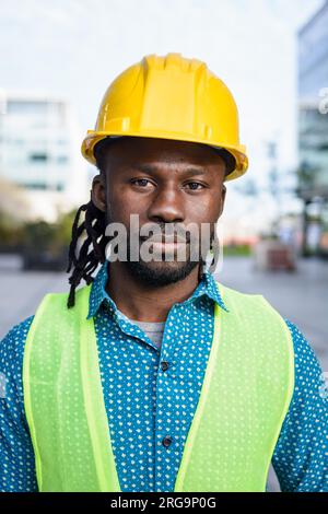 Vertikales Porträt eines schwarzen Ingenieurs mit kurzem Bart und Dreadlocks. Er trägt einen Helm und eine reflektierende Weste und steht draußen und schaut sich das an Stockfoto