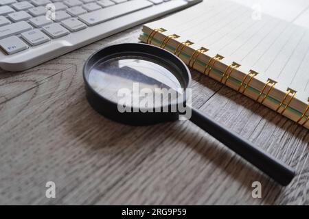 Nahaufnahme von Vergrößerungsglas, Notebook und Computertastatur auf dem Tisch. Konzept recherchieren, untersuchen und untersuchen Stockfoto