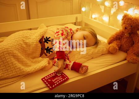 Ein bezauberndes Kleinkind, das mit Teddybär schläft, und ein Brief an den Weihnachtsmann in der Hand unter dem Weihnachtsbaum. Ein Kind träumt Silvester im Bett. Stockfoto