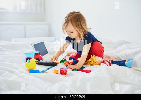 Ein kleines Mädchen mit Laptop, Notebook, Telefon und verschiedenen Spielzeugen im Bett auf sauberer weißer Bettwäsche. Freiberufler, Fernunterricht oder Arbeit von zu Hause mit Kindern CO Stockfoto