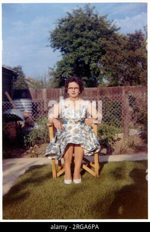 Eine Frau mittleren Alters in einem floralen Sommerkleid, die in einem Gartenstuhl aus Holz und Leinwand in einem gepflegten Vorstadtgarten sitzt. Stockfoto