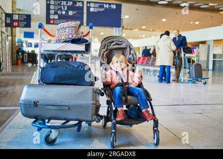 Ein bezauberndes kleines Kleinkind auf dem internationalen Flughafen. Kleines Kind, das in einem Kinderwagen in der Nähe von schwerem Gepäck sitzt. Auslandsreisen mit Kindern Stockfoto