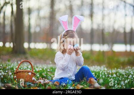 Ein Mädchen mit Hasenohren, das Ostern auf Eierjagd spielt. Das Kleinkind sitzt auf dem Rasen mit vielen Schneetropfen und sammelt bunte Eier im Korb. Beleuchtet Stockfoto