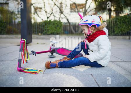 Vorschulmädchen im Einhorn-Helm, das nach dem Sturz auf dem Boden saß, während sie an einem Frühlingstag im Park mit dem Roller fuhr. Outdoor-Sportaktivitäten und Stockfoto