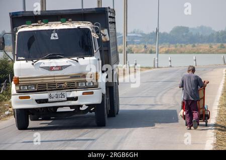 SAMUT PRAKAN, THAILAND, FEBRUAR 01 2023, Ein Truck fährt neben einem alten Mann, der einen Trolley auf einer Landstraße schiebt Stockfoto