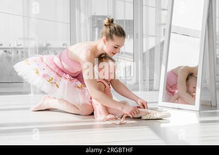 Zwei Ballerinas, Mutter und Tochter, die im Studio vor großen Fenstern auf dem Garn sitzen. Die kleine Ballerina und ihre Lehrerin machen die Teilung. Ein kleines Mädchen, das sich mit seiner Mutter streckt Stockfoto