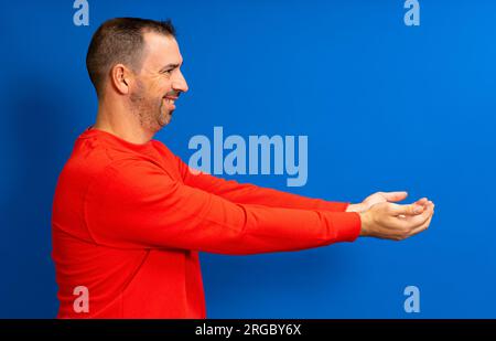 Bärtiger hispanischer Mann in seinen 40s Jahren trug einen roten Pullover und bot etwas mit ausgestreckten Händen an, isoliert auf blauem Studiohintergrund. Stockfoto